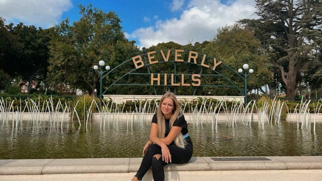 Die glückliche Gewinnerin unsere Mitarbeiter-Gewinnspiels sitzt vor einem Brunnen und einem "Beverly Hills"-Schild in Los Angeles.