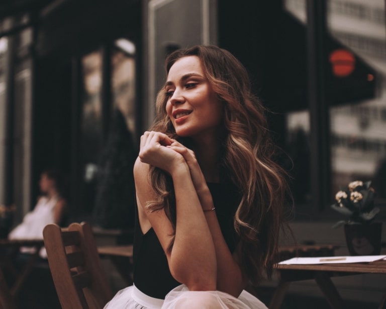 Une femme aux longs cheveux bruns balayés est assise sur une chaise dans un café et regarde vers la droite.